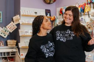 Two girls where matching black sweaters that read "Self love club"