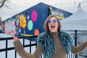 Caroline Stokes poses in front of the Grindstone Comedy Bistro. She is wearing sunglasses and a green and blue fur vest.