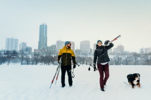 A couple cross country ski with their dog. The downtown skyline is in the background.