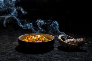 vegetables in a large bowl, with a bundle of sage burning beside it. The smoke from the sage is visible hovering over the bowl of food.