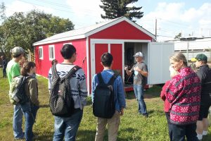 A man gives a tour to a group of people at the Edmonton Urban Farm in front of a red shed.