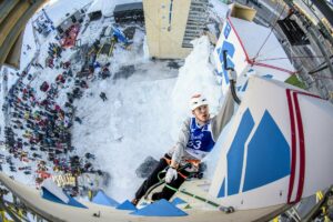 A man climbing an ice wall with a crowd watching in the background