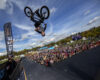 A man is seen upside down on his BMX bike after hitting a half pipe jump. There is a crowd of people behind him in the background an a FISE World flag.