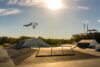 A BMX rider is seen soaring in the sky across a skatepark with his hands on the BMX bike. The sun is shinning down on him and a blue sky with green trees are in the background