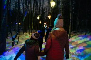 A mother holds her daughters hand while walking through a colourfully lit forest trail at night.