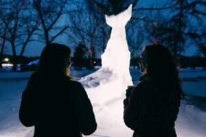 Two people face a lit snow sculpture with a spotlight on it during evening.