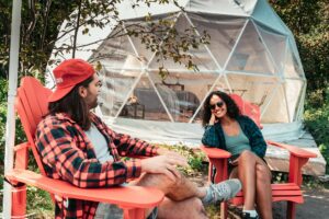 A couple sits in some red chairs outside their geodesic glamping domes on a summer day.
