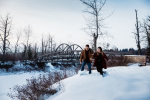 A couple run through the snow with a bridge in the background.