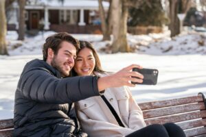 A couple takes a selfie on a bench on a sunny winter day.