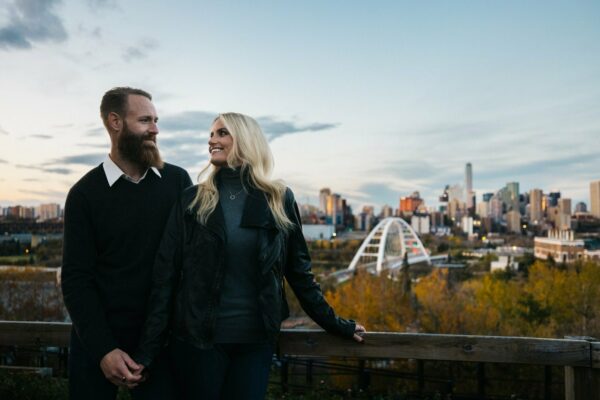 A couple smiles and poses in front of a view of the Walterdale Bridge and Edmonton city skyline.